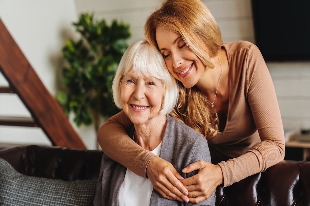 misconceptions about assisted living Happy daughter embracing from behind elderly mother at living room. Love and care. Family time and bonding concept. Assistance and support of elder generation