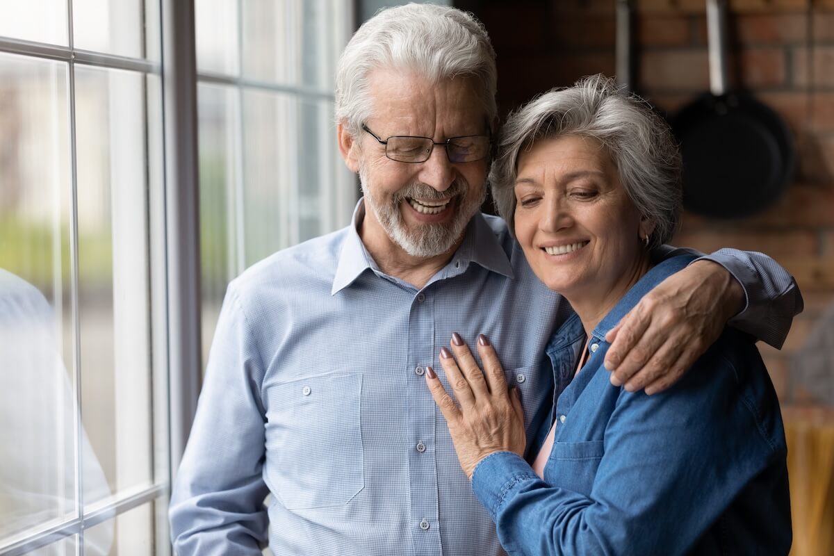 Independent Living Benefits | Astral at Auburn Happy old mature hoary man in eyeglasses cuddling smiling beautiful middle aged wife, daydreaming together at home near window. Loving older senior family couple enjoying tender sweet moment indoors.
